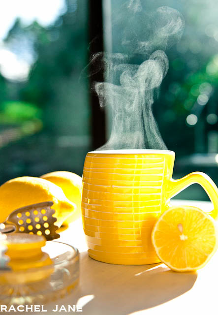 White table with a large yellow mug and steam coming out of the top with large yellow lemon on the side. 